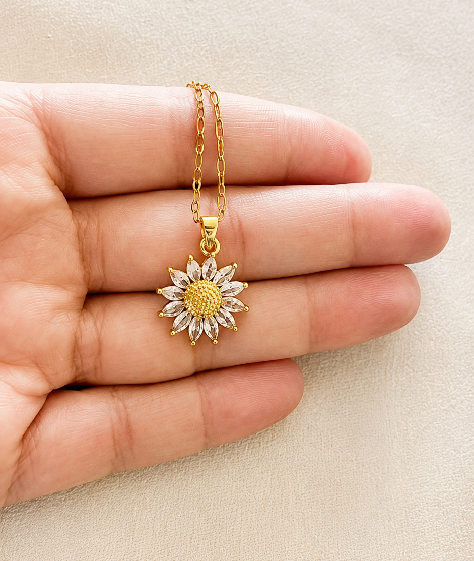 Gold necklace with a sunflower pendant held between fingers on a beige background