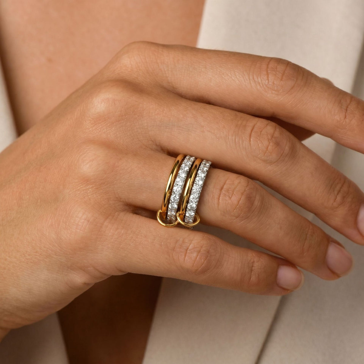 A close-up of a hand wearing three slim gold rings, including two pavé-set diamond bands, against a soft beige background.