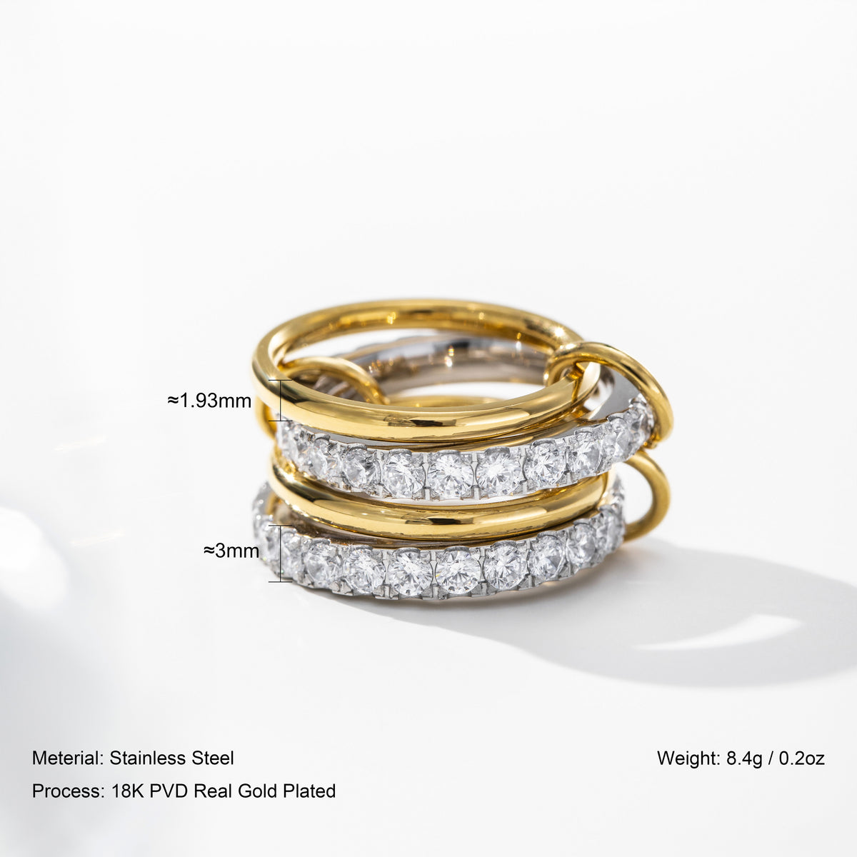 A close-up studio shot of a stack of three rings on a white background. The top ring is plain yellow gold, the middle ring is gold with a row of clear stones, and the bottom ring is silver-toned with a row of clear stones.