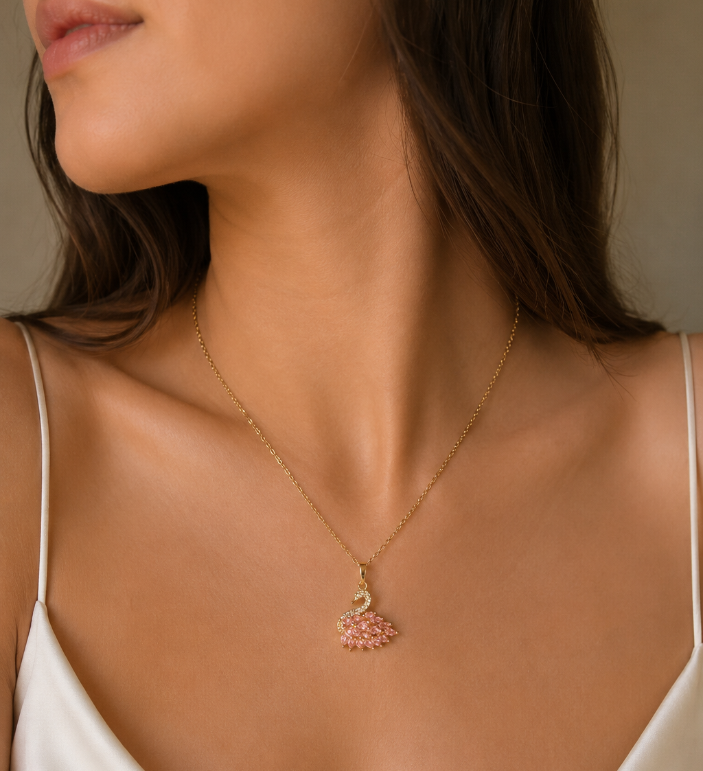 A close-up lifestyle shot of a model wearing a delicate gold chain necklace with a small pink and gold swan pendant, styled with a white satin camisole top.