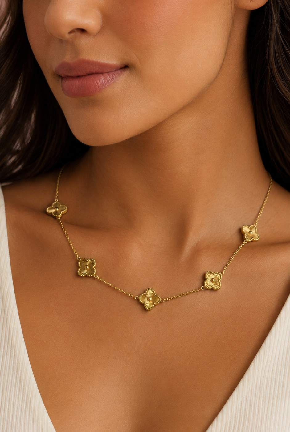 A close-up of a woman wearing a delicate gold chain necklace with five small clover-shaped charms, styled with a white ribbed V-neck top.
