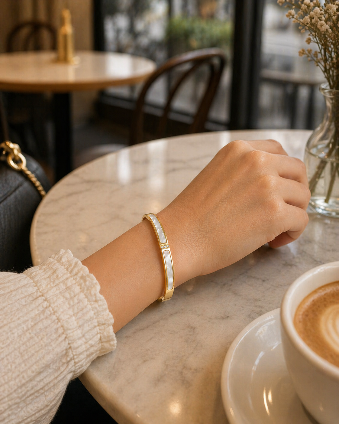 Person's hand wearing a gold bracelet on a marble table with a cup of coffee.