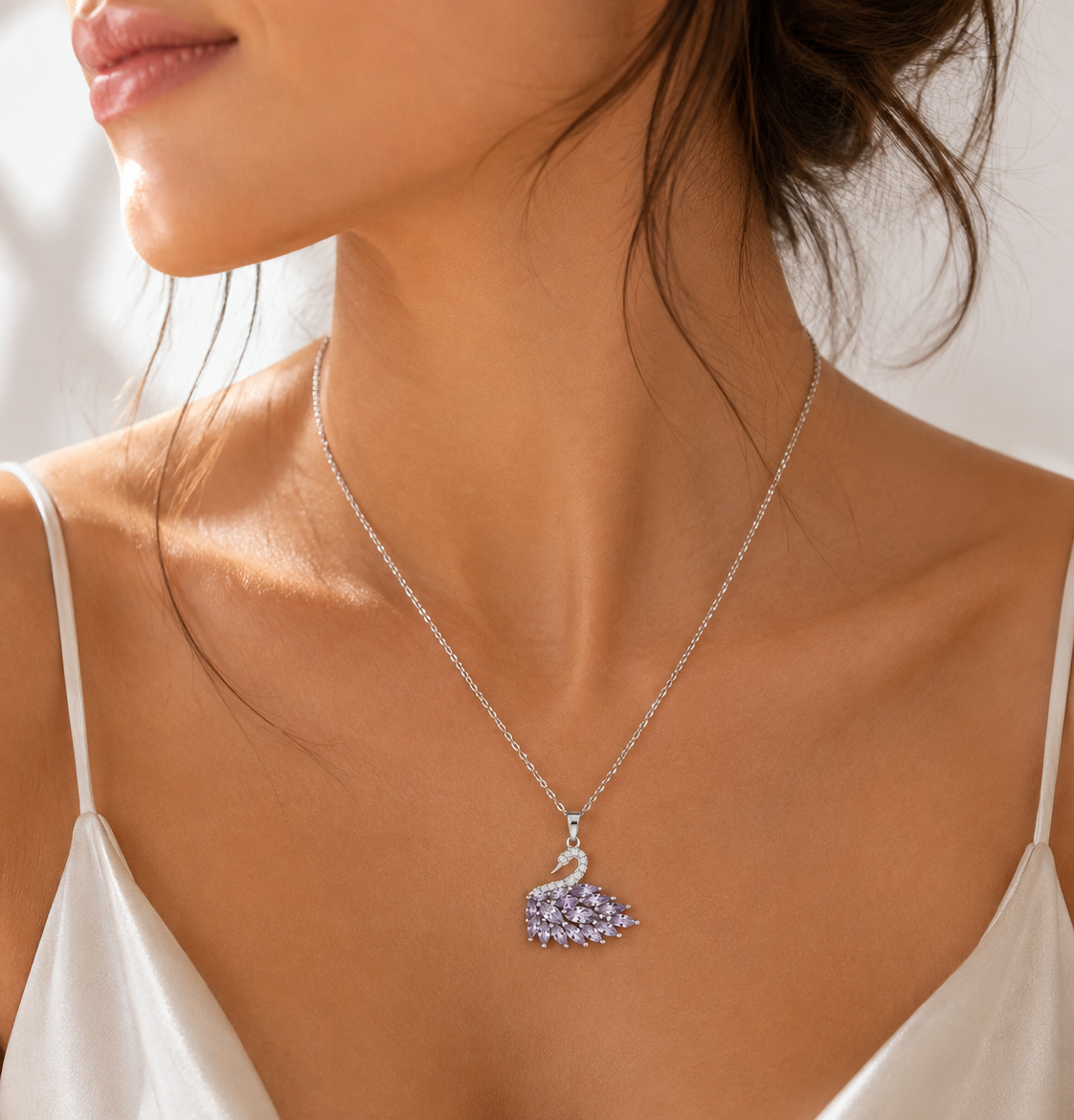 A close-up lifestyle shot of a model wearing a delicate silver chain necklace with a crystal swan pendant, styled with a white satin camisole.