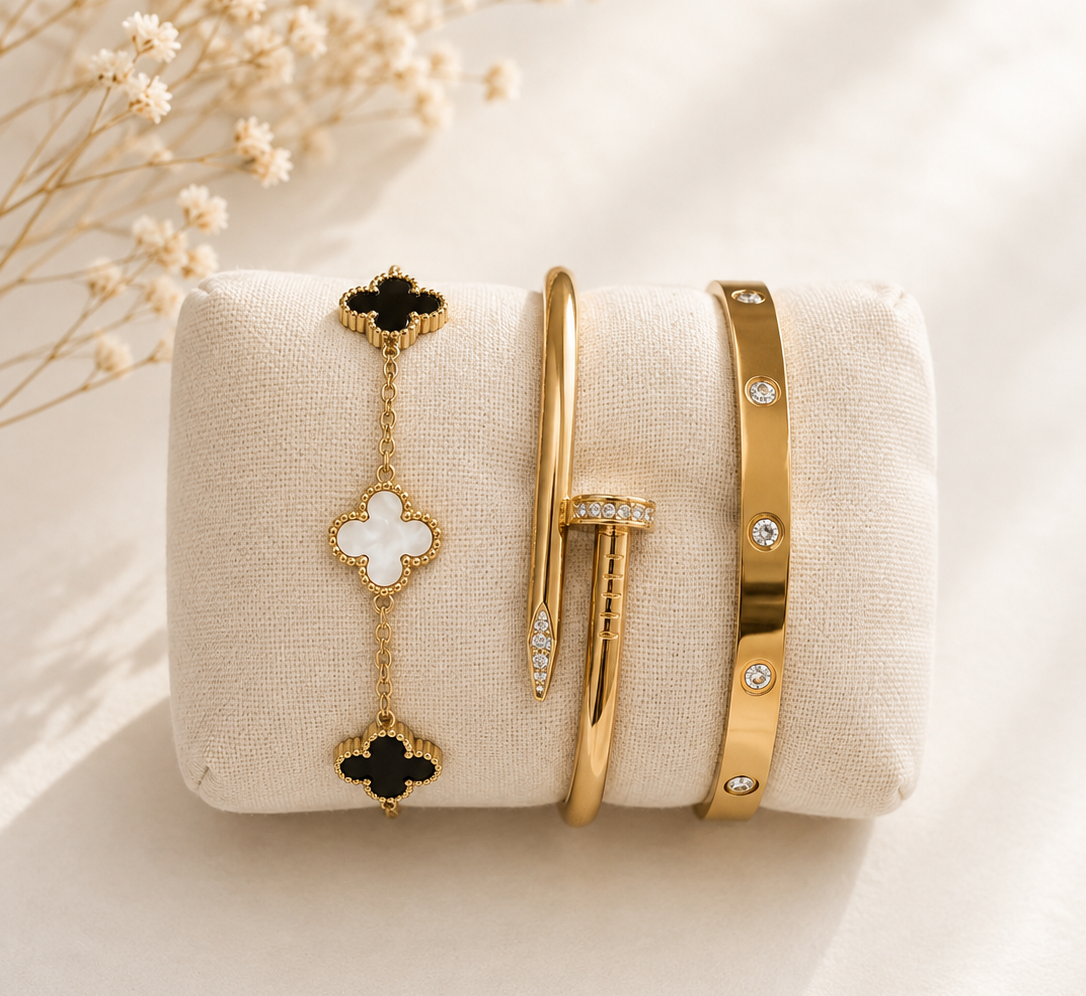 A set of gold-toned jewelry bracelets displayed on a cream fabric jewelry stand, with dried floral stems in the upper left corner. The set includes a bracelet with black and white quatrefoil motifs, a slim gold bangle with a pavé-set stone, and a gold bangle with small clear rhinestone accents.