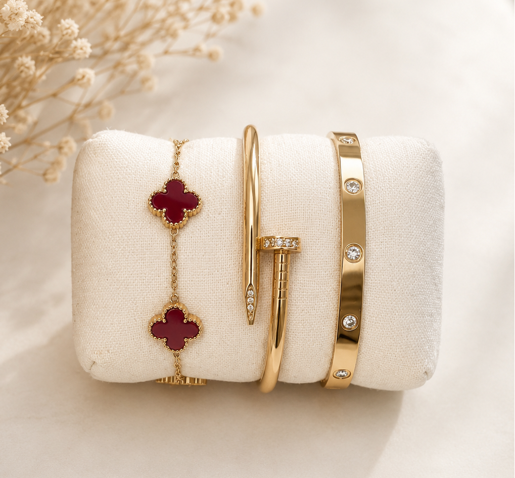 A set of gold jewelry displayed on a cream-colored velvet jewelry stand, featuring a delicate gold chain bracelet with two red clover motifs, a slim gold bangle with a pavé diamond tip, a gold bar with pavé diamonds, and a gold bangle with diamond accents. The stand is styled with dried baby's breath flowers on a light marble surface.