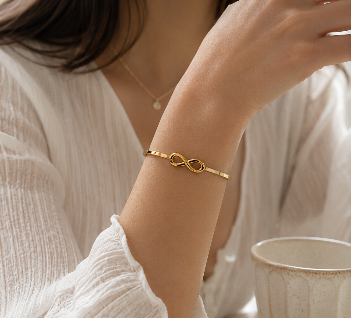 A gold infinity bracelet worn on a woman's wrist, shown in a lifestyle setting with a white blouse and a ceramic mug. The lower portion features a text block titled 'INFINITY BANGLE' with product description and feature icons.