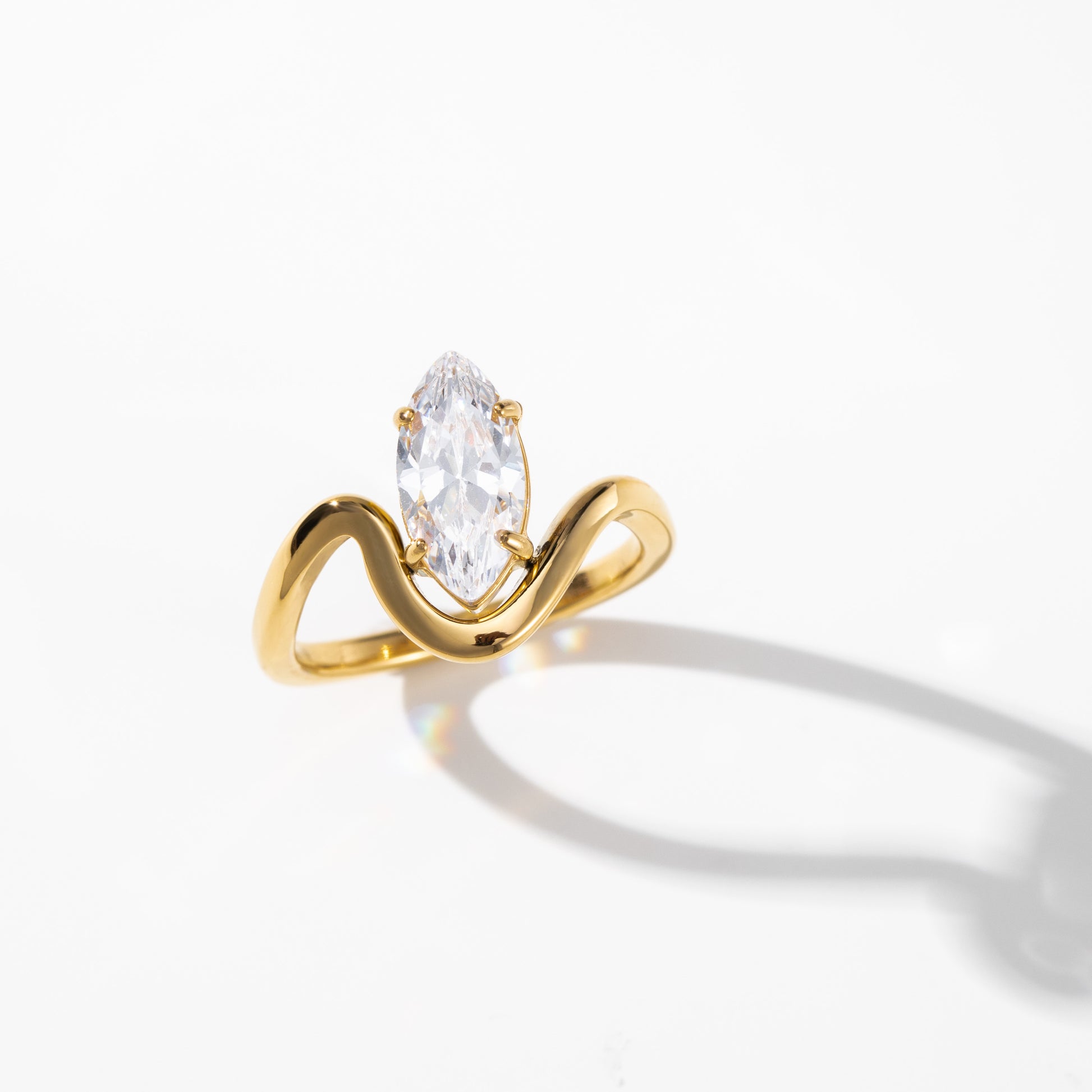 A yellow gold ring featuring a marquise-cut clear gemstone in a curved, wave-like setting, photographed on a clean white background with a soft shadow.