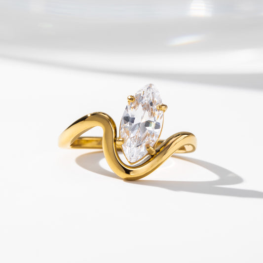 A gold ring with a marquise-cut clear gemstone, set in a wavy, flowing band design, photographed on a clean white surface with soft shadows.