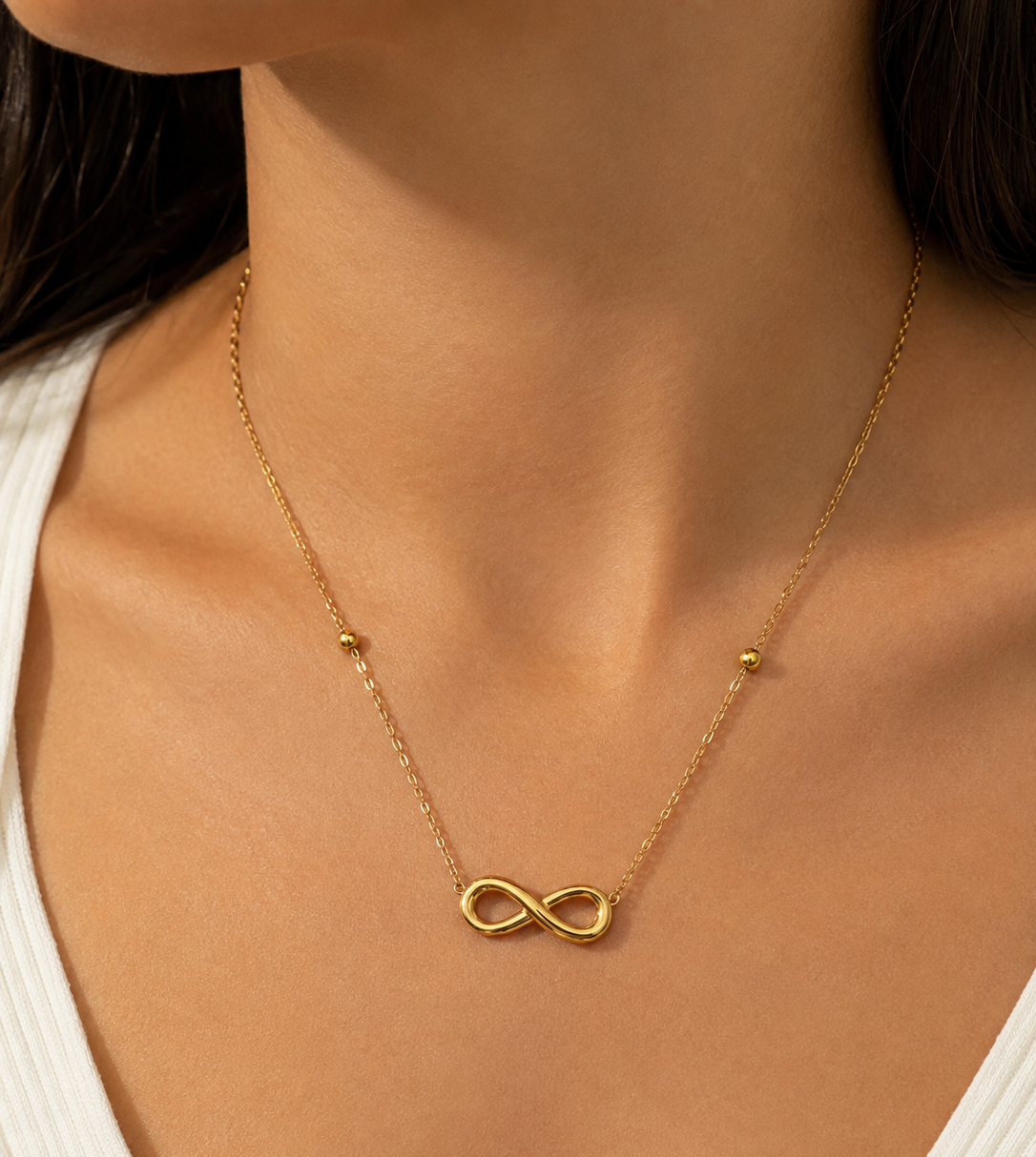 A close-up of a model wearing a delicate gold chain necklace with a small infinity symbol pendant and tiny gold bead accents on the chain.