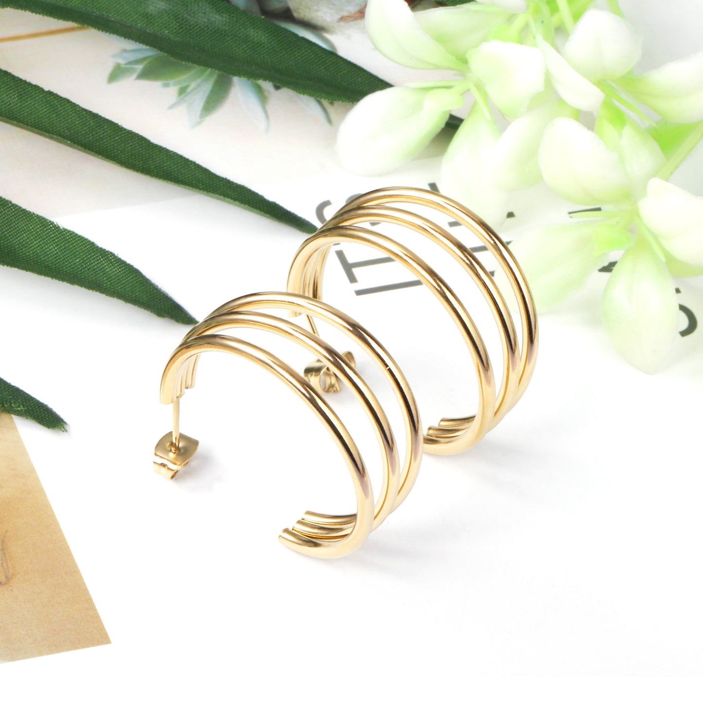 Gold hoop earrings on a white surface with green leaves and flowers in the background